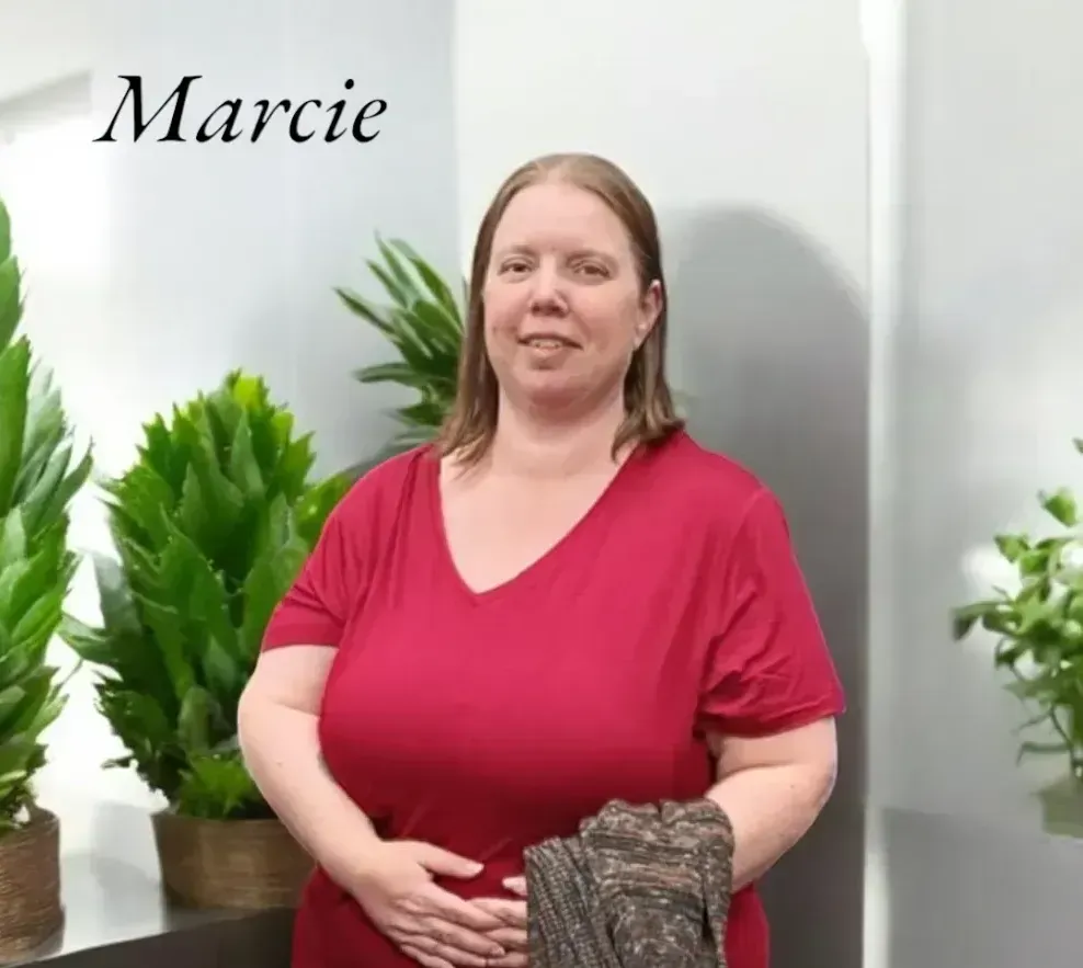 A person with shoulder-length hair wearing a red v-neck shirt stands in front of a white wall with potted plants.