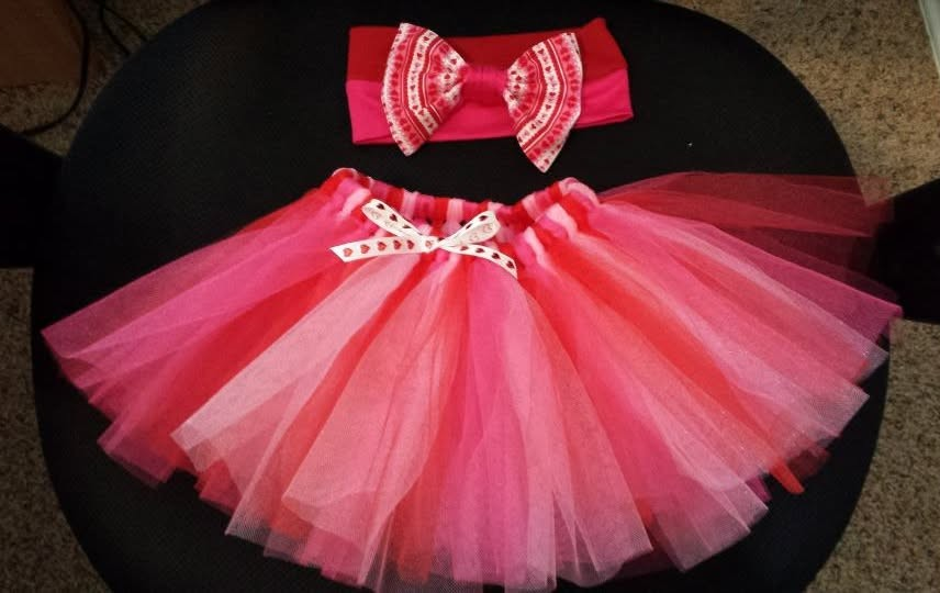 A red and pink tulle tutu with a matching headband featuring a patterned bow, laid on a dark surface.