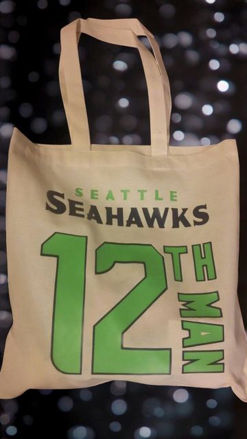 A Seattle Seahawks "12th Man" tote bag in beige with dark green and lime green text, set against a blurry bokeh background.