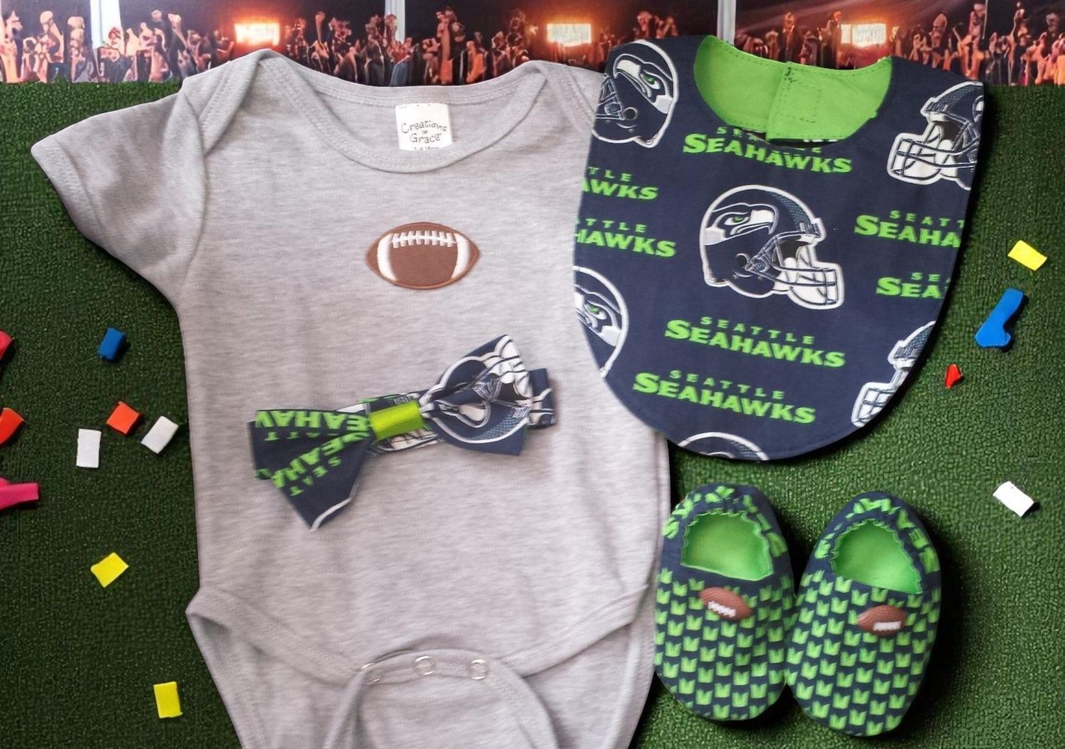 A grey baby bodysuit with a football, a Seahawks print bib, a matching bow tie, and booties on a green artificial turf.