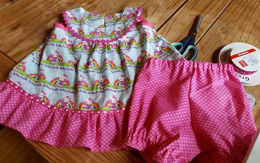 A floral and unicorn print toddler dress with matching pink patterned bloomers on a wooden surface with sewing scissors.