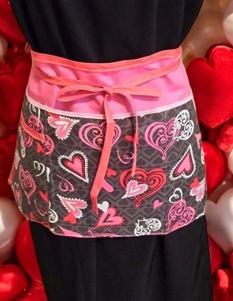 A waist apron featuring a black fabric with various pink and white heart patterns, tied with a bright pink waistband.