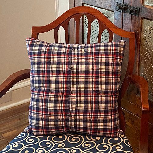 A square pillow made from a plaid flannel shirt, featuring a button-down placket, resting on a decorative wooden chair.