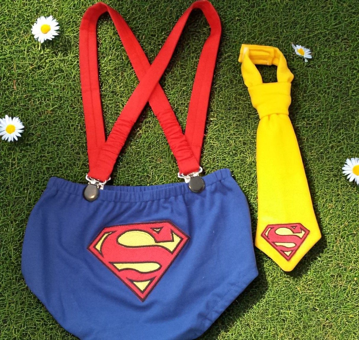 Blue Superman-themed baby diaper cover with red suspenders and a matching yellow necktie, placed on green grass.