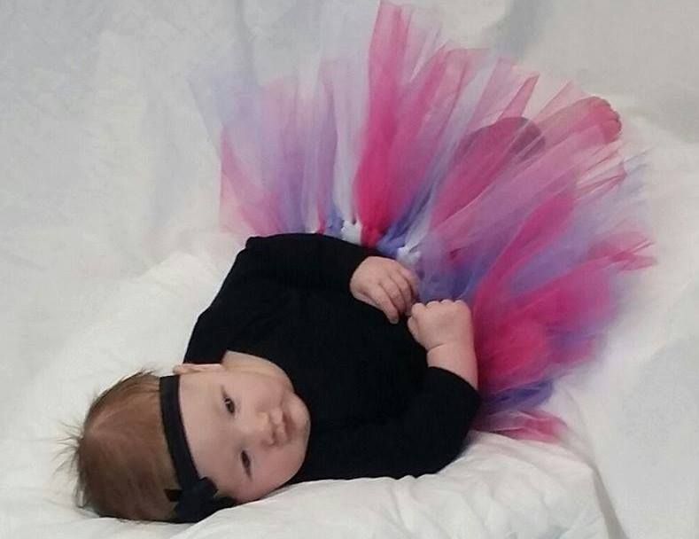 A baby wearing a black long-sleeved shirt and a pink, blue, and white tulle tutu lies on a white surface.