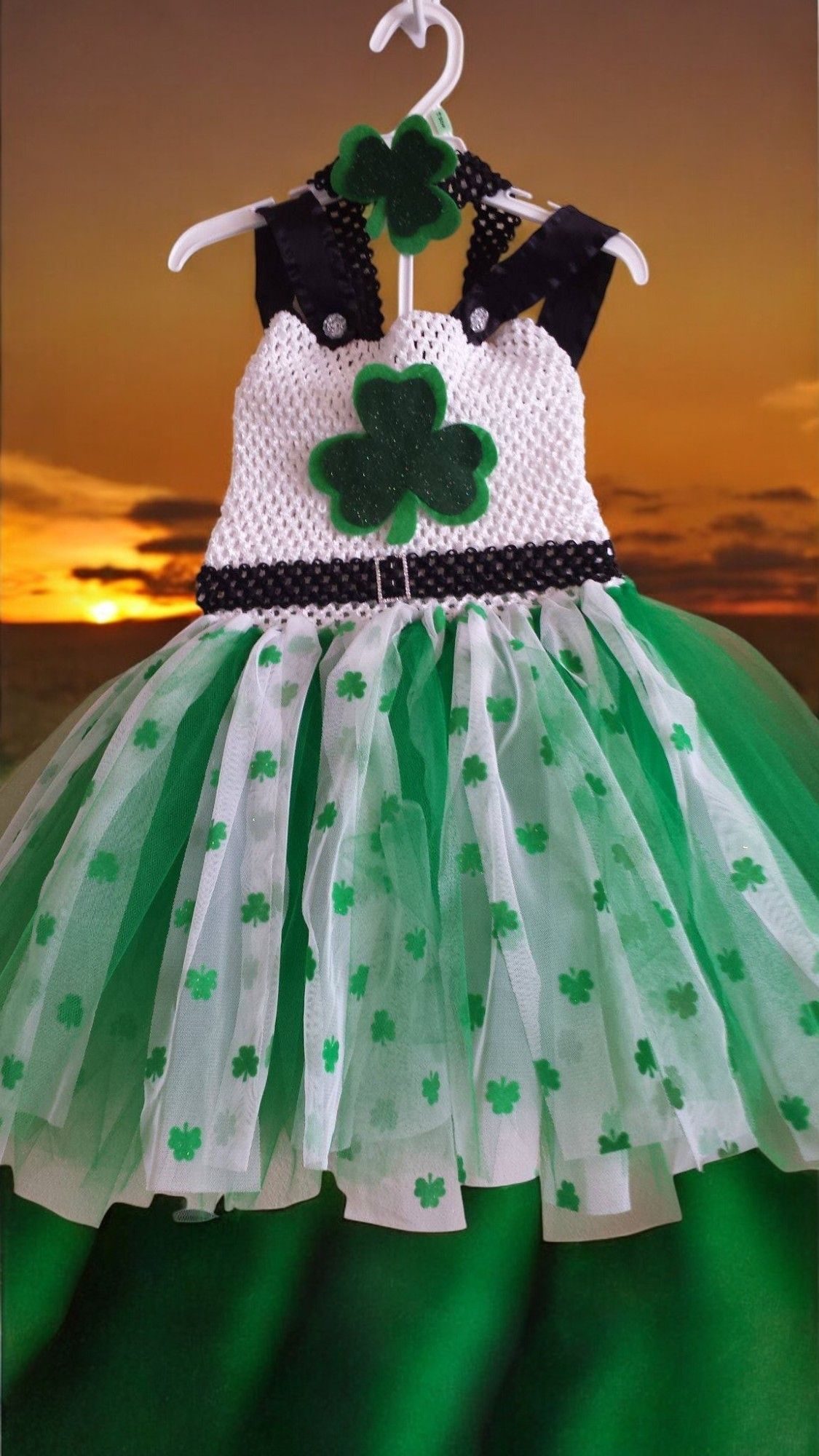 A St. Patrick's Day tutu dress with a white crochet top, clover accents, and green tulle skirt, hung on a hanger.