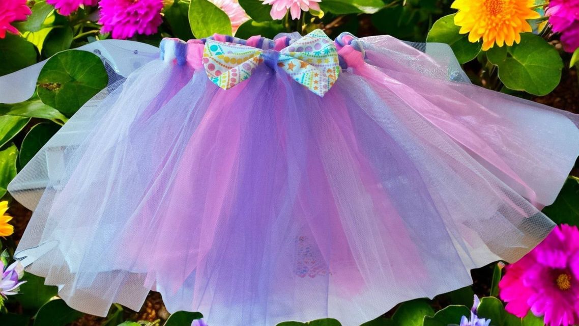 A multi-colored tulle tutu in shades of purple, blue, and pink, featuring a decorative floral bow, set against greenery.