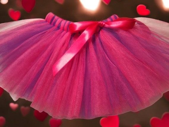 A fluffy, layered tutu in shades of pink and purple, featuring a bright pink satin bow on a dark background with hearts.