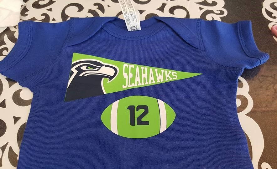 Blue baby onesie with a green Seattle Seahawks pennant graphic and a football icon featuring the number 12.