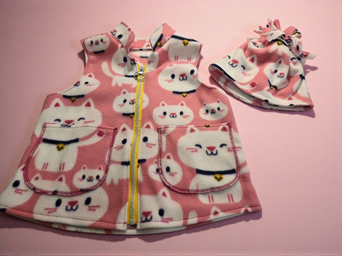 A pink vest and matching hat with a white cat print, featuring a gold zipper and two pockets, set against a pink backdrop.