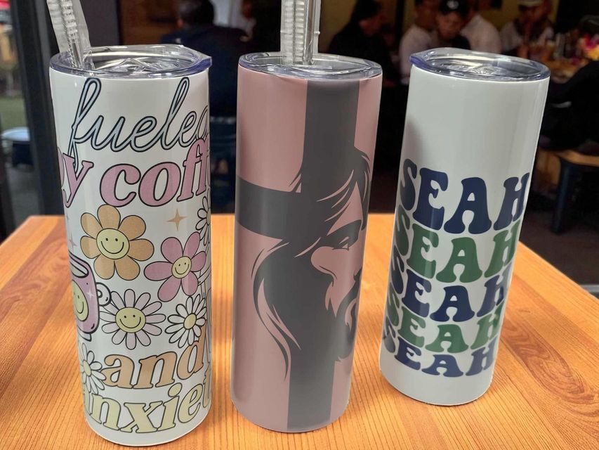 Three tumblers with straws stand on a wooden table: one with flower patterns, one with a portrait, one with text.