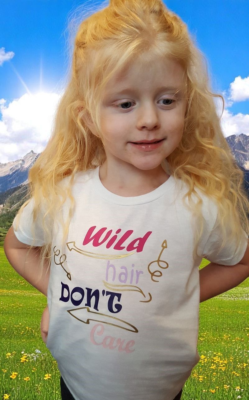A young child with wavy blonde hair stands in a field with mountains in the background, wearing a shirt that says 