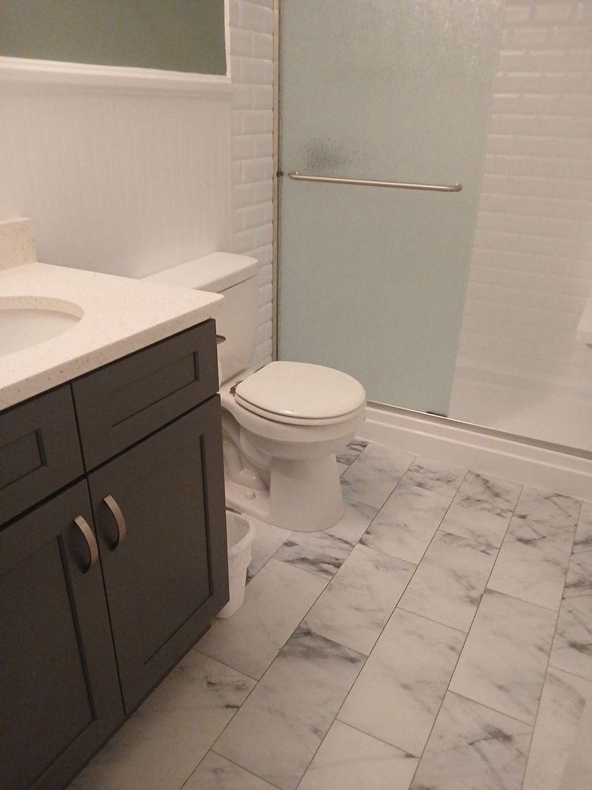 Modern bathroom with dark vanity, white sink, toilet, marble tile floor, and frosted shower door