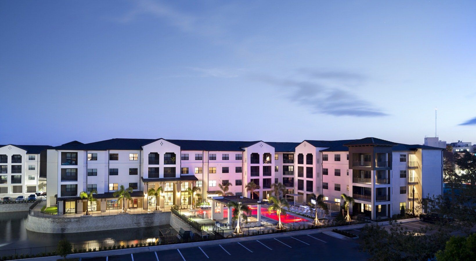 Apartment complex at dusk with illuminated pool, water feature, and blue sky.