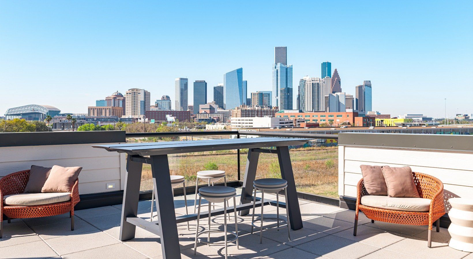 Rooftop patio with a city skyline view; table with stools, wicker chairs, and pillows on a sunny day.