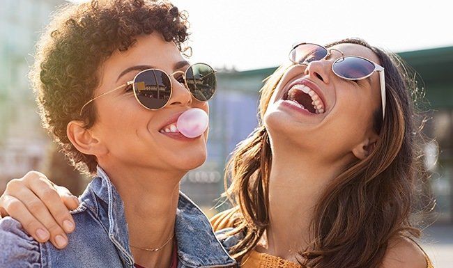 Two smiling women in sunglasses, one with a bubblegum bubble, outside.