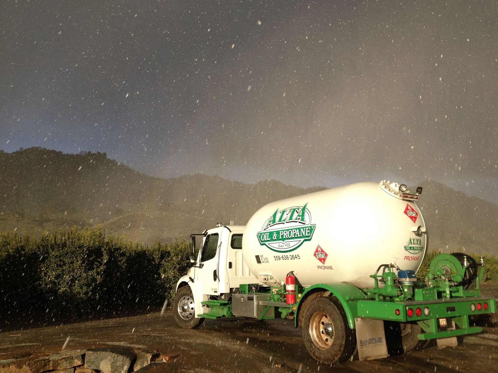 White truck with green accents parked near bushes, with a mountain background in the rain.