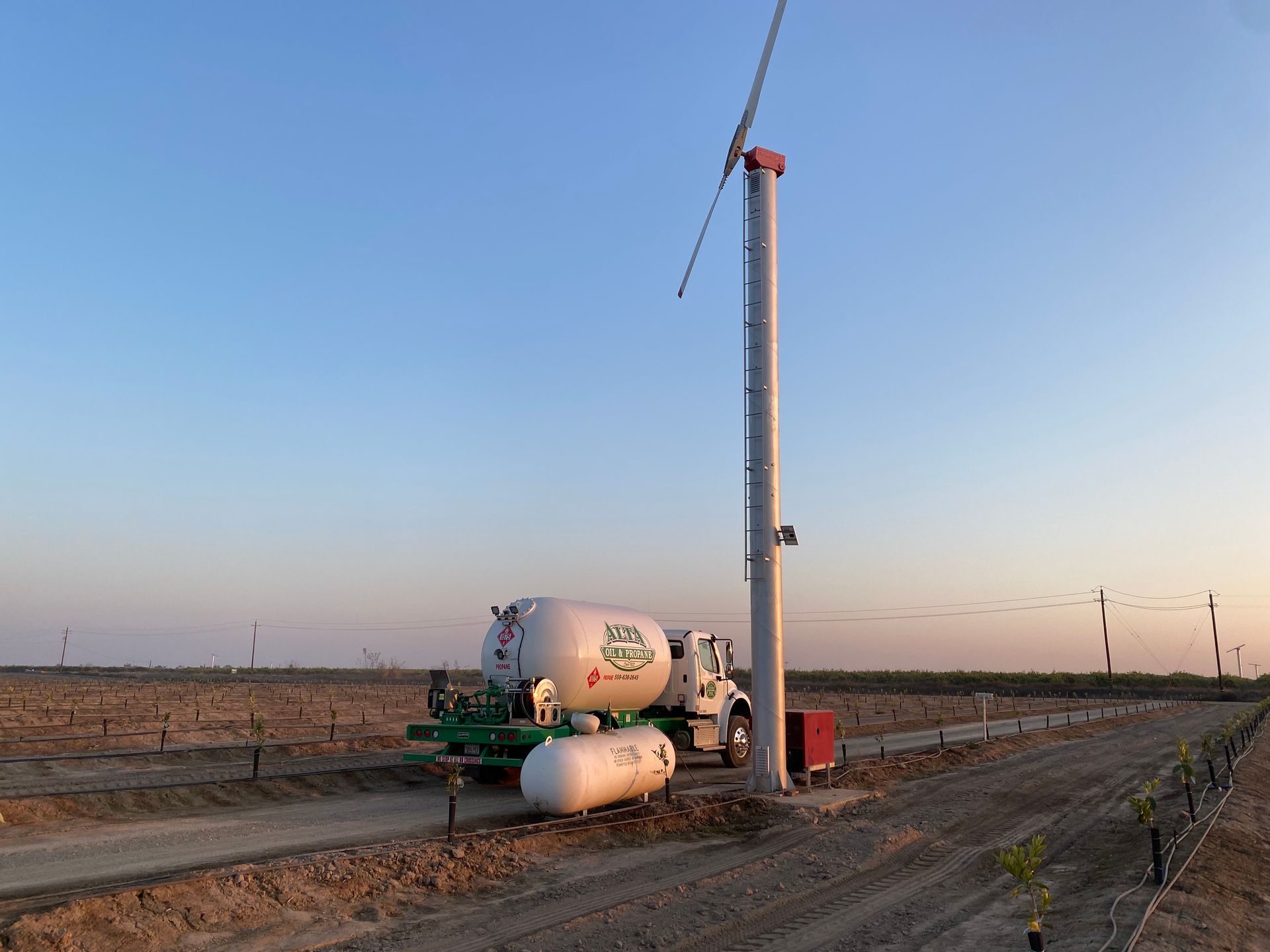 A truck with a large white tank beside a tall tower in a field, under a blue sky.