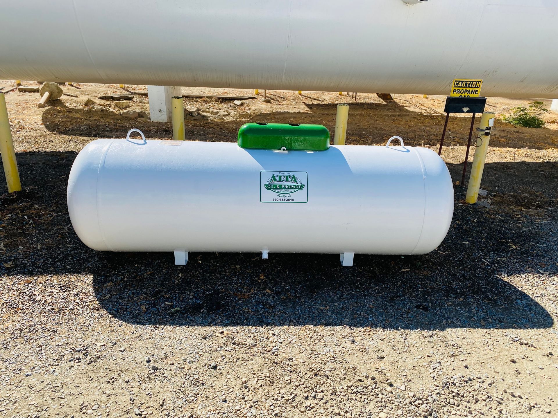 White propane tank with green top and logo, under a white pipe on gravel.