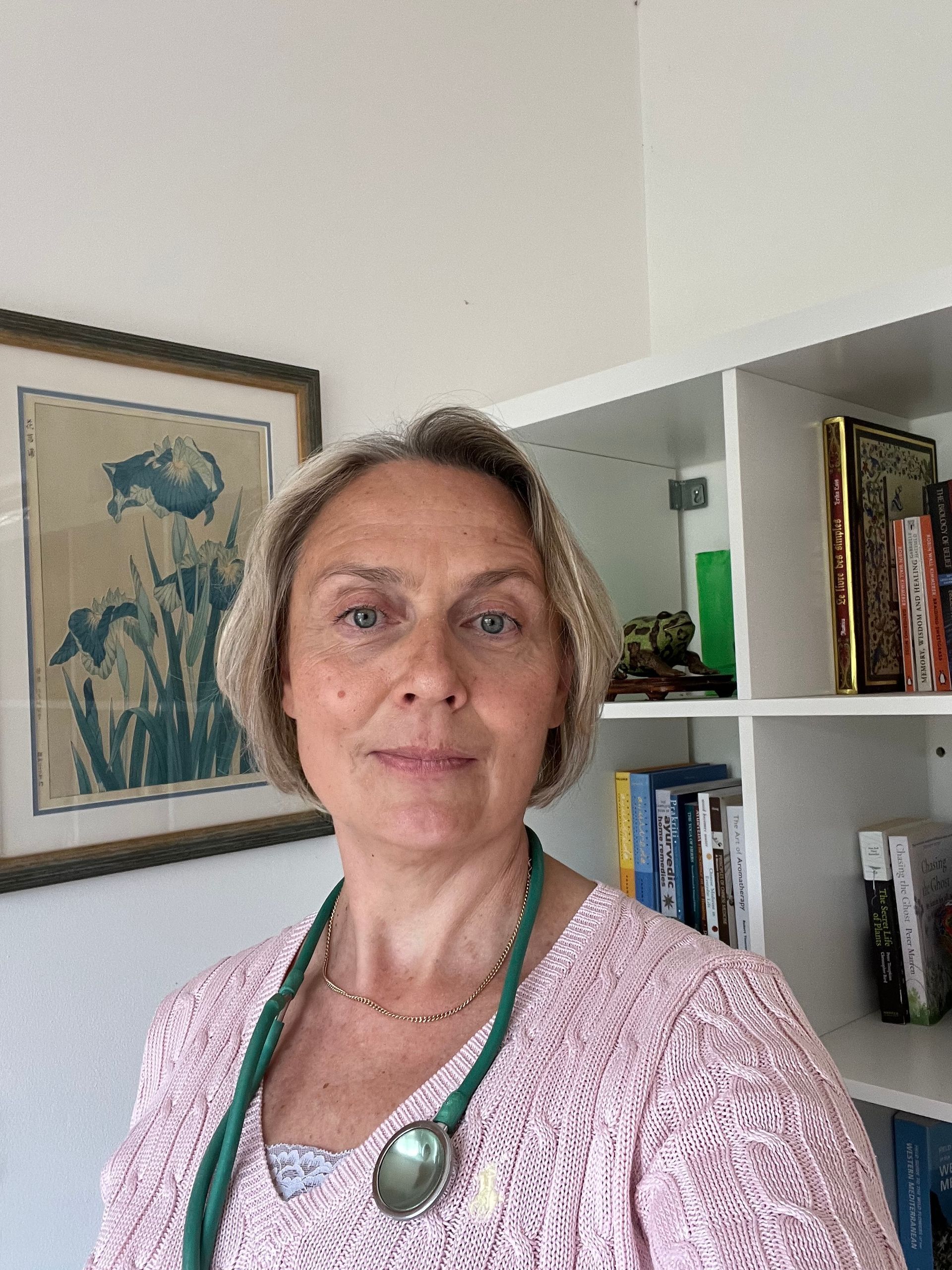 Woman standing indoors in a pink top, with framed artwork and a bookshelf behind her.