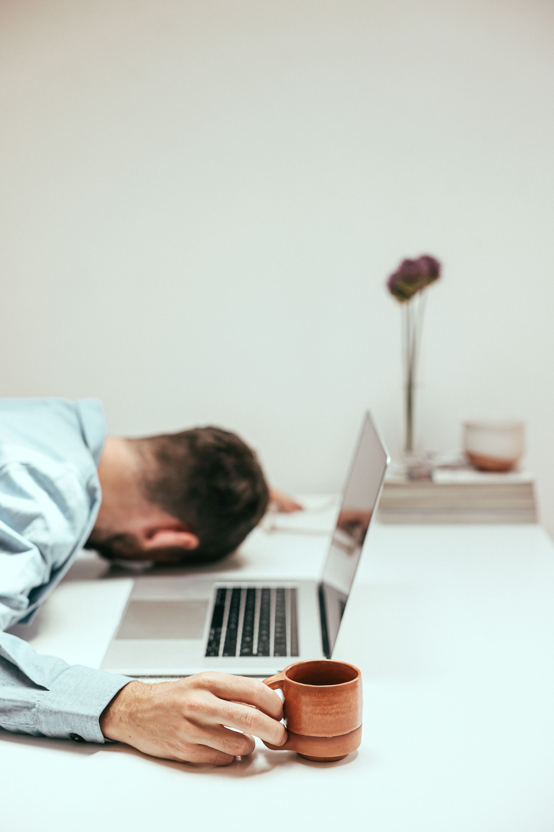 Office worker sleeping face-down at a desk beside an open laptop and coffee mug