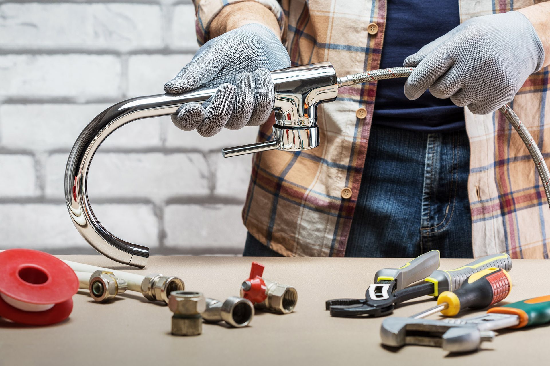 Plumber in gloves, fixing a faucet, surrounded by tools and parts, against a brick wall background.