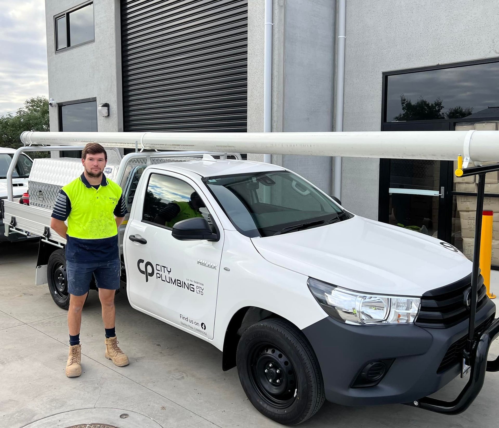 Man in work clothes stands beside a white truck carrying long pipes. Building is in the background.
