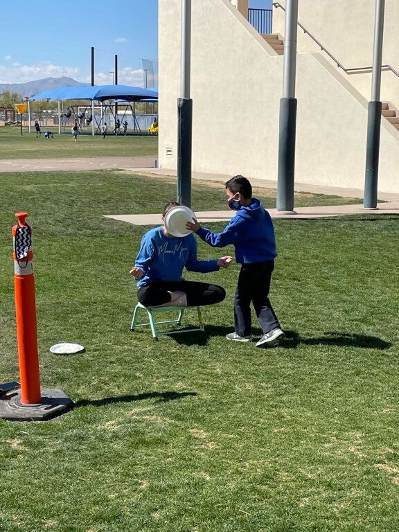 Two people in blue shirts kneel on grass near a building, with a child touching a seated person’s hat.