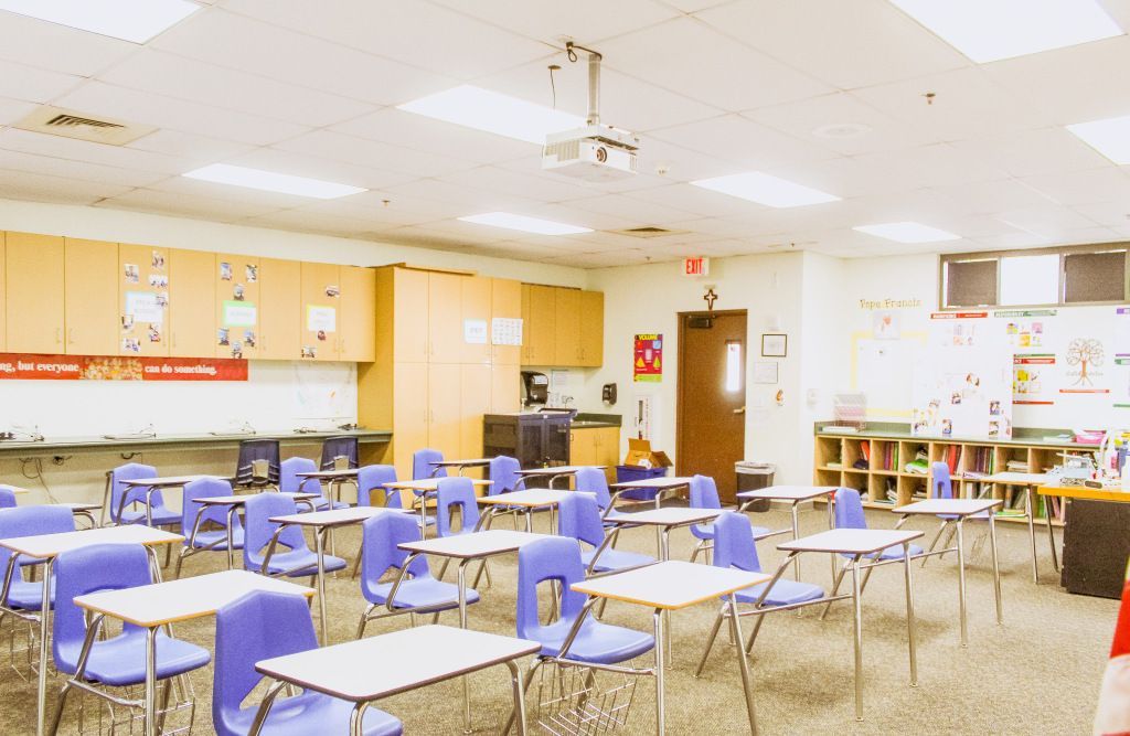 Empty classroom with blue desks, white chairs, and wood cabinets under bright ceiling lights.
