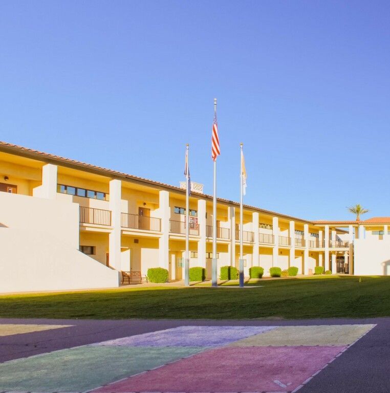 Low-rise hotel building with balconies and flags under a clear blue sky, seen from a landscaped lawn.