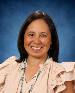 Smiling woman in a beige blouse and beaded necklace against a blue studio backdrop
