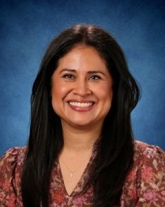 Smiling person with long dark hair in a patterned blouse against a blue studio background