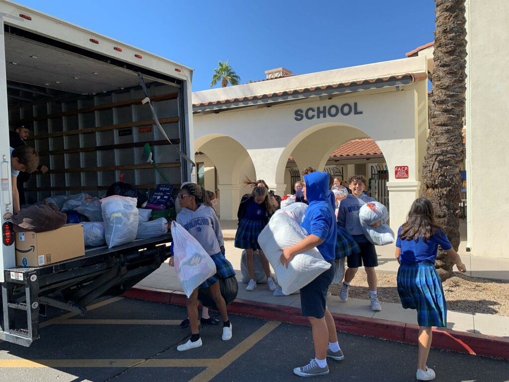 Volunteers unload donated bags from a truck outside a school building with an arched entrance.