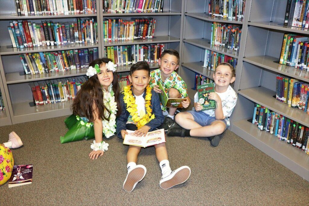 Four children sitting on the floor in a library aisle, smiling with books around them.