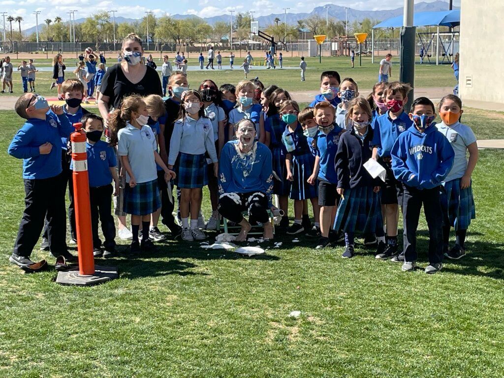 A group of children in school uniforms pose on a grassy field with an adult and a bright orange cone.