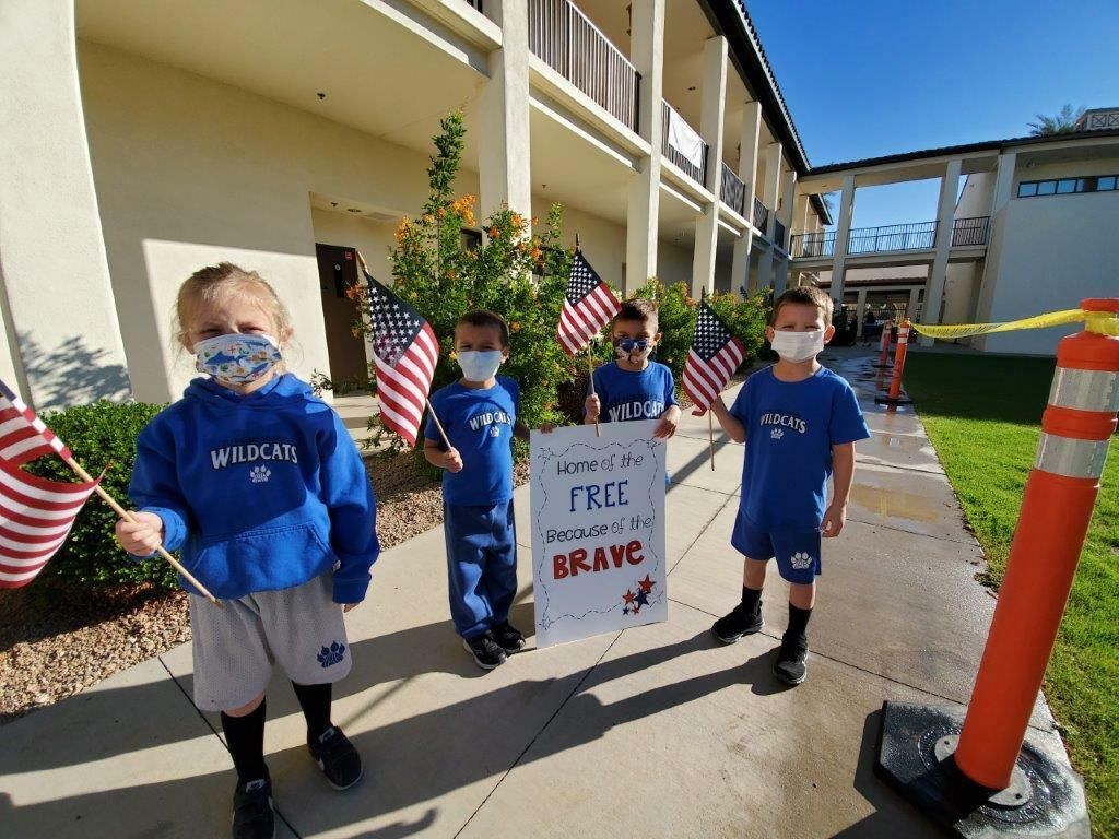 Masked students in blue shirts hold a sign outside a school building, with U.S. flags and an orange cone nearby.