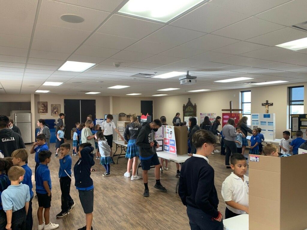 Children gather in a bright cafeteria, standing in lines and groups around tables during an indoor event.