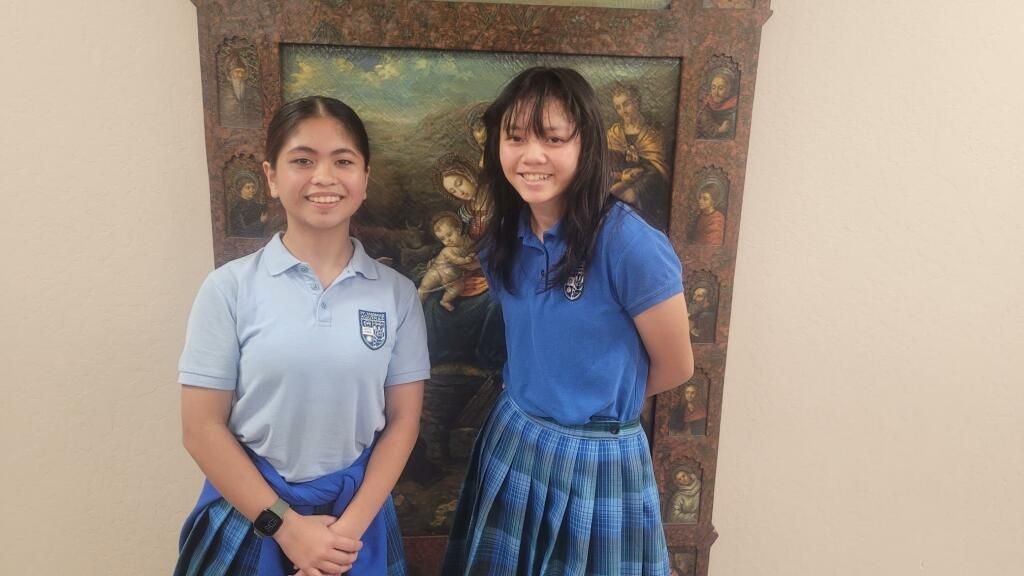 Two students stand smiling in front of a framed painting indoors.