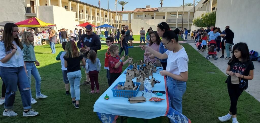 People gathered outdoors around display tables in a grassy courtyard, browsing handmade crafts and projects.