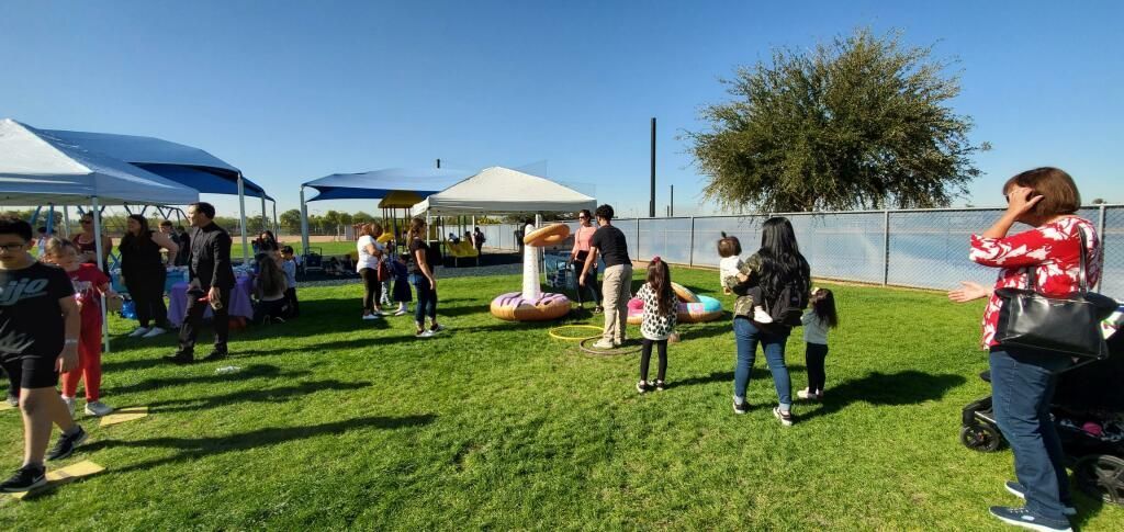 People gathered on a grassy field near white tents and a tree on a sunny day.