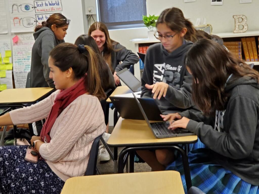 Students collaborating at desks in a classroom, using laptops and talking together.