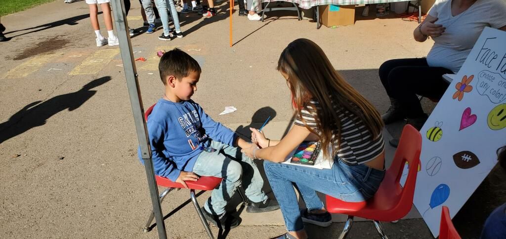 Child and woman seated outdoors at a microphone during a small event, with a colorful sign nearby