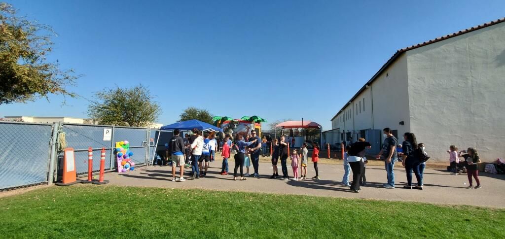 People gathered outside a long white building on a sunny day, with orange cones and grassy foreground.