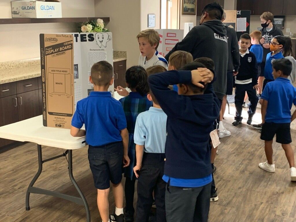 Children in blue shirts crowd around a display board at an indoor event.