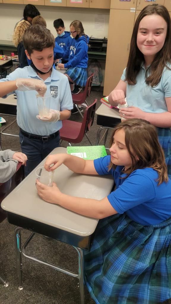 Students in a classroom; one pours liquid into a cup while others watch nearby.