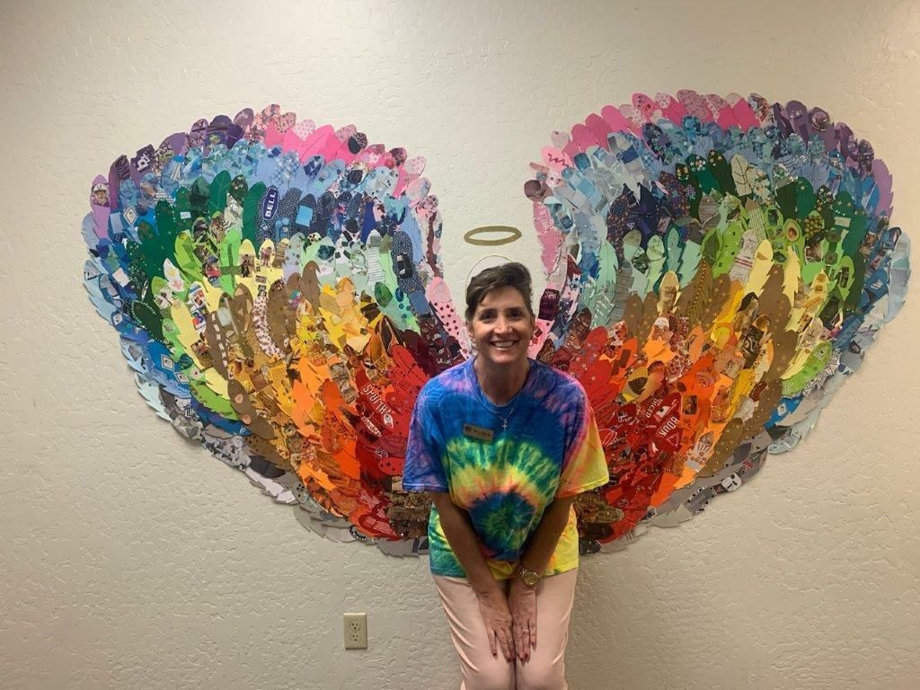 Person kneels before colorful butterfly wing mural, smiling in a rainbow tie-dye shirt.