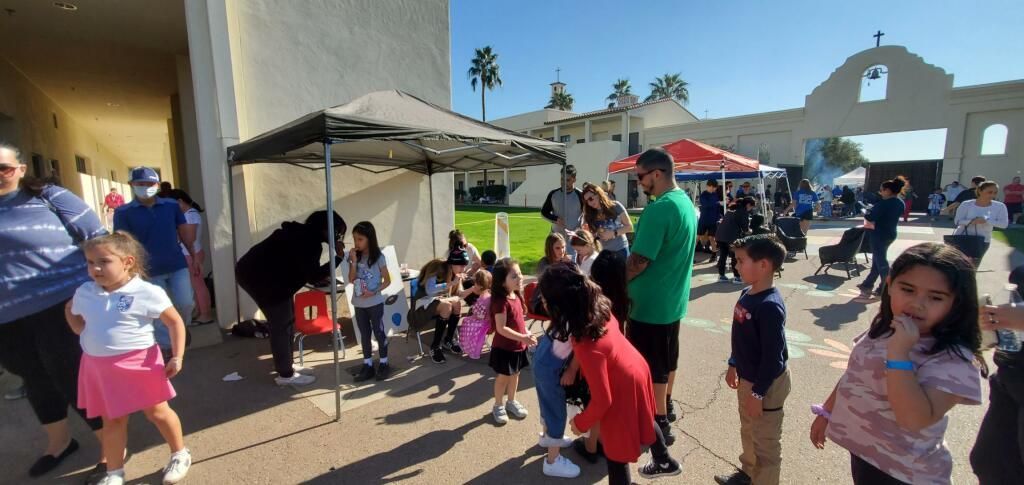 People gathered in a sunny school courtyard, children playing near a canopy tent and adults mingling nearby.