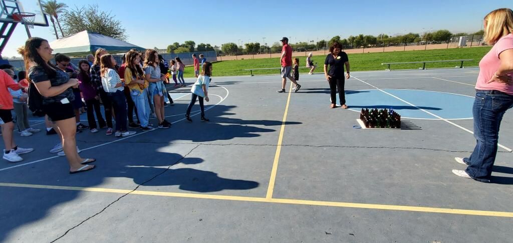 Students gathered on a school parking lot beside a blue-painted court, watching an activity on the pavement