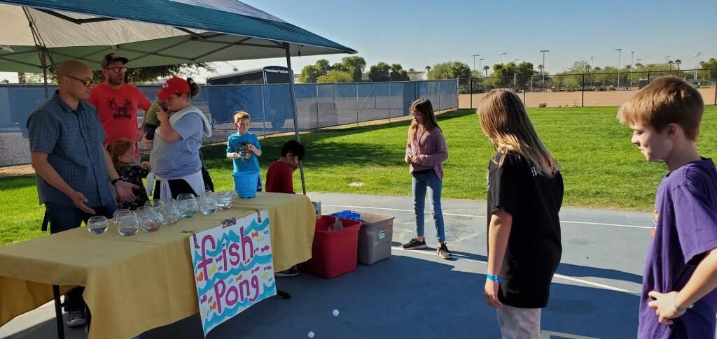 People at a colorful outdoor booth under a blue canopy, with a sign and table in a sunny field.