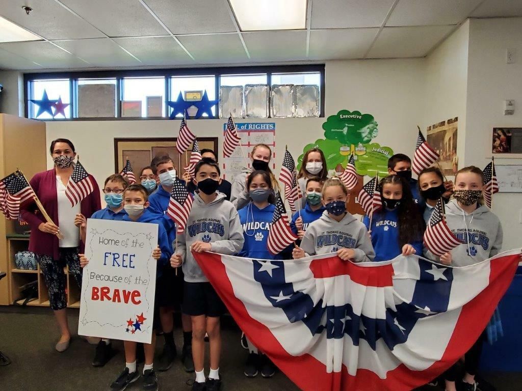 Students hold American flags and patriotic signs in a classroom decorated for a U.S. celebration.